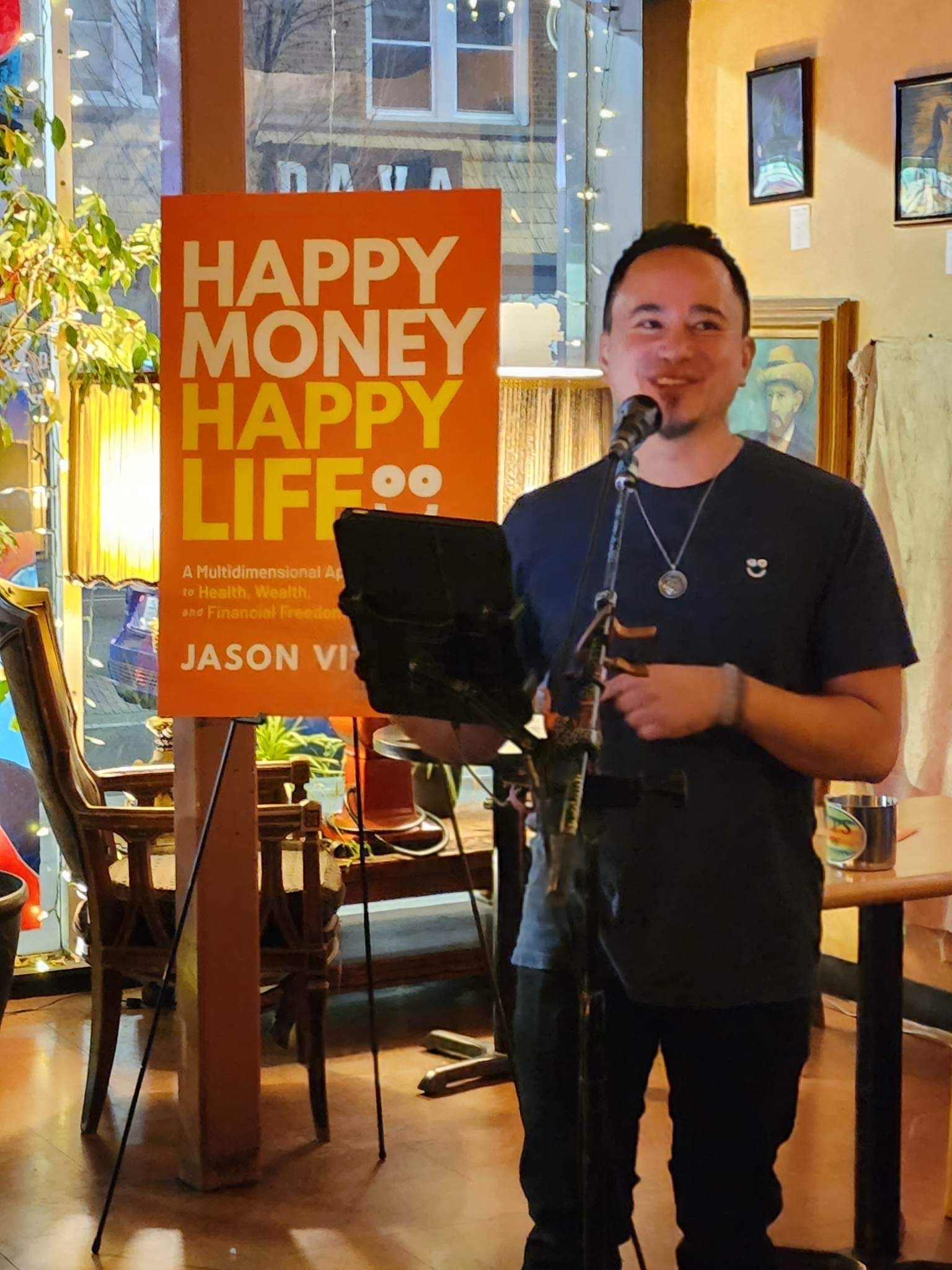 photo of author Jason Vitug reading an excerpt of his book, 'Happy Money Happy Life' which is prominently featured on a poster behind him.
